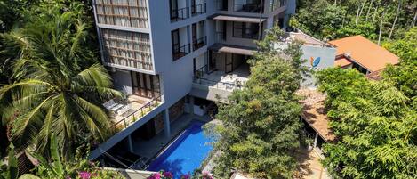A multi-story building with balconies, a swimming pool, and lush greenery.
