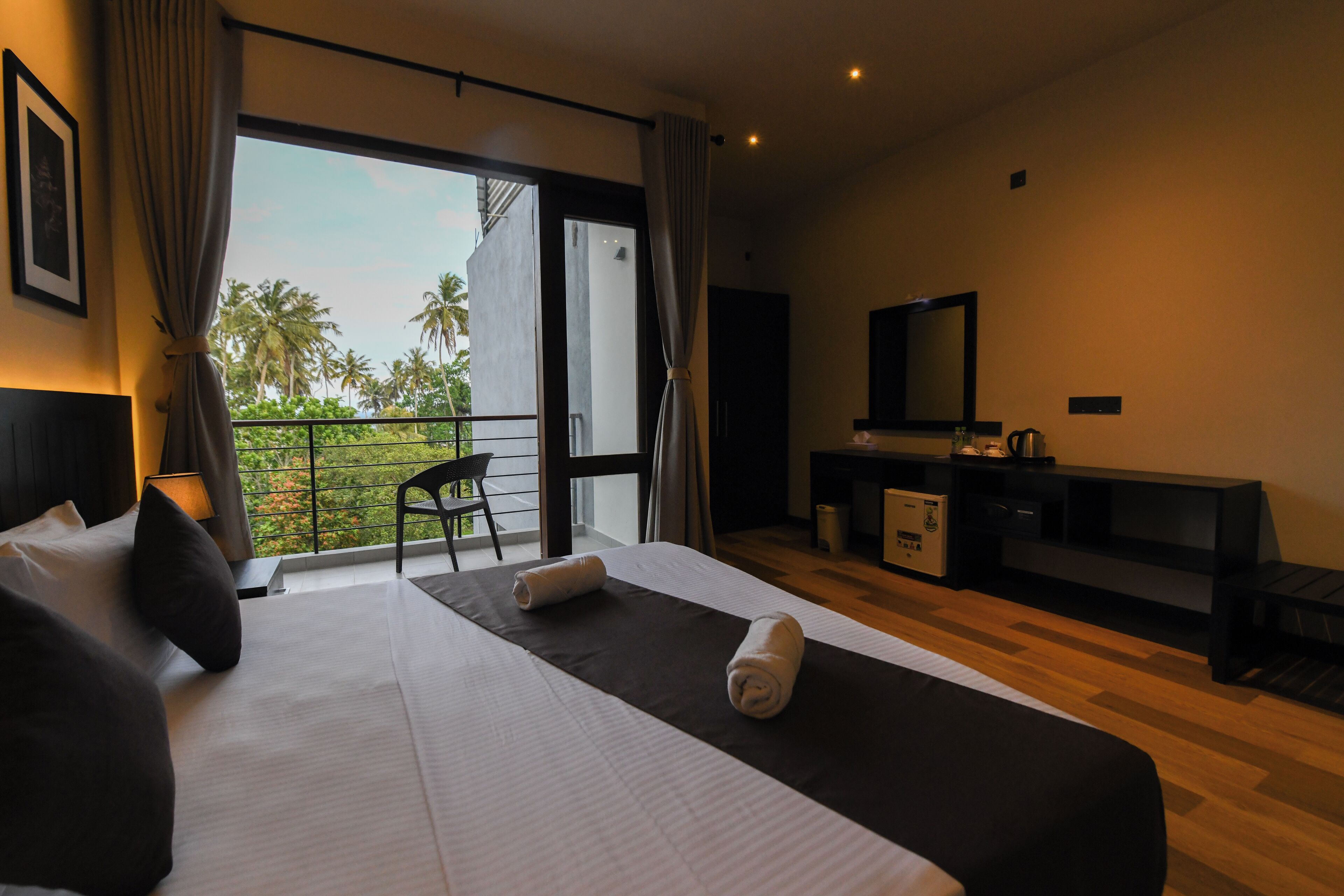 Habitación de hotel con una cama grande, un escritorio, una silla y un balcón con vistas a palmeras.