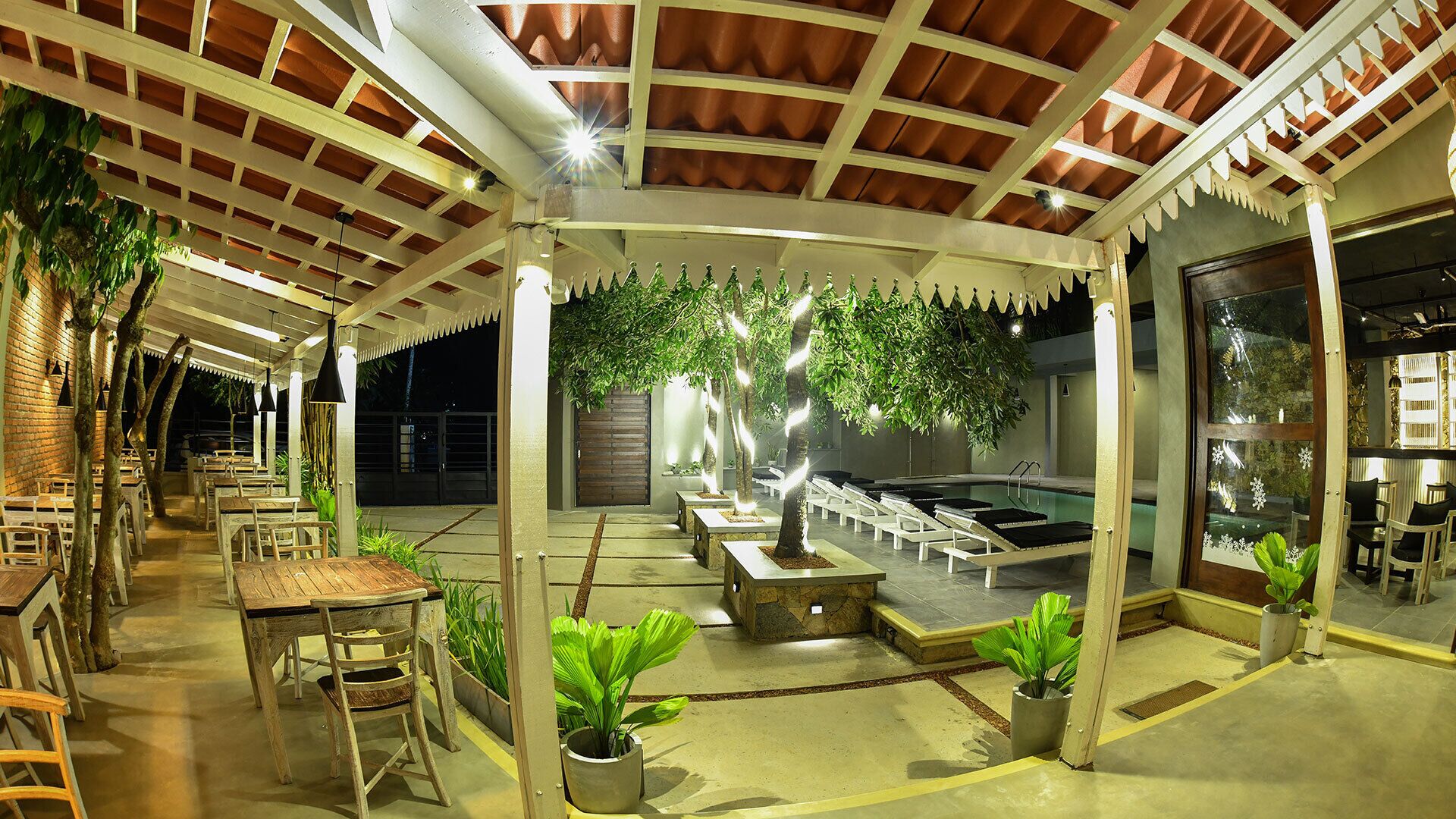 An outdoor dining area with wooden tables and chairs, a pool, and potted plants.
