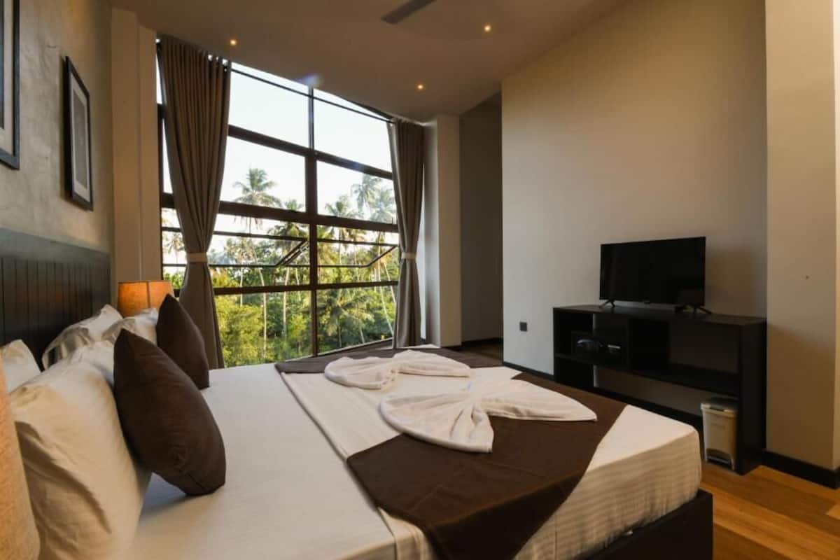 Habitació d'hotel amb una finestra gran, un llit amb almoades, un televisor i un petit armari.