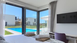Een moderne hotelkamer met een groot bed, een bureau, een stoel en een televisie. Er is een zwembad zichtbaar door de ramen.