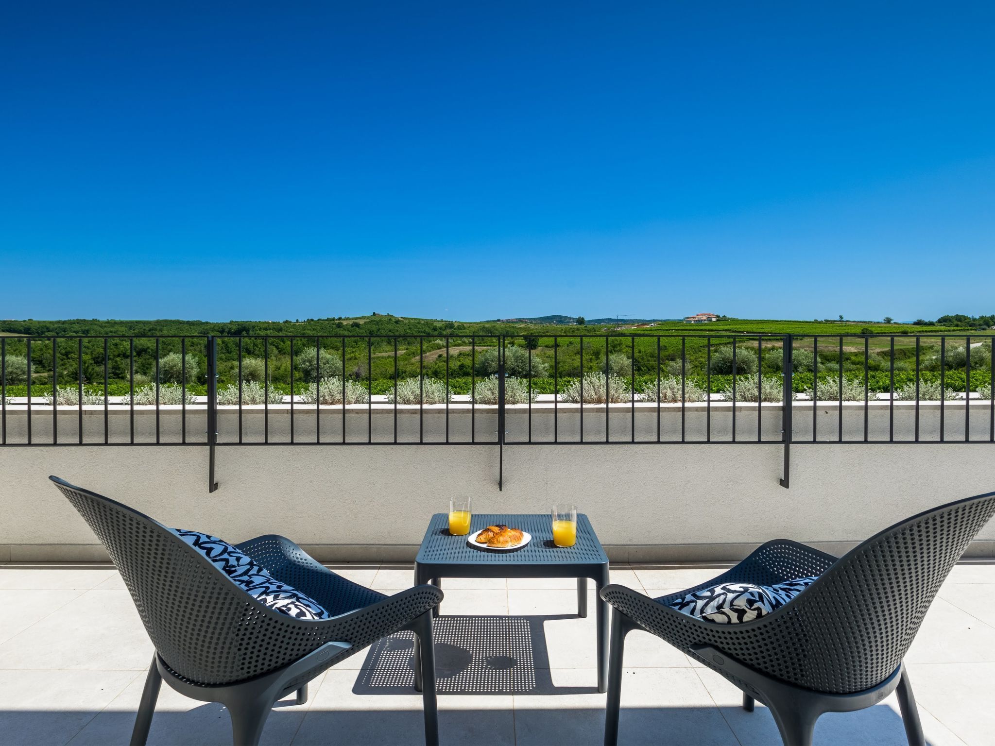 A balcony with two chairs and a table, a clear view of a green landscape, and a metal railing.