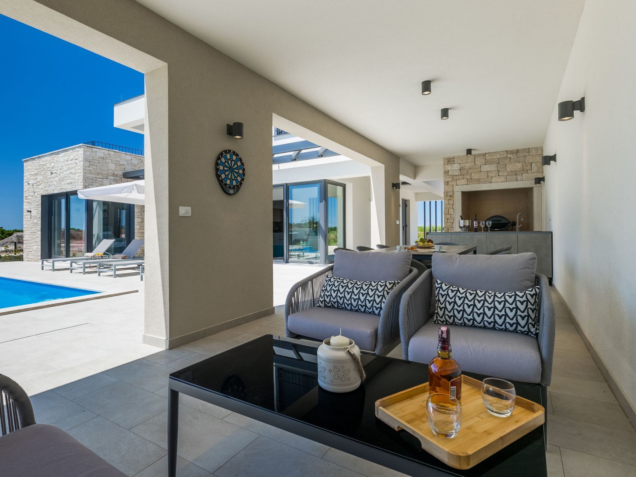 A modern living room with a pool view, a dartboard, and a dining area with a stone fireplace.