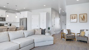 A modern living room with a sectional sofa, two armchairs, a dining area with wicker chairs, and a kitchen with white cabinets and a refrigerator.
