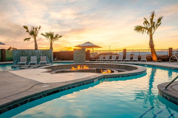 Piscina con sillones de descanso y una chimenea, rodeada de palmeras y vista al atardecer.