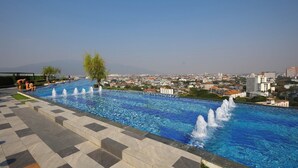 A swimming pool with water fountains, surrounded by a tiled deck, with a cityscape in the background. - Luxury Astra Sky River Apartment (Chang Wat Chiang Mai)