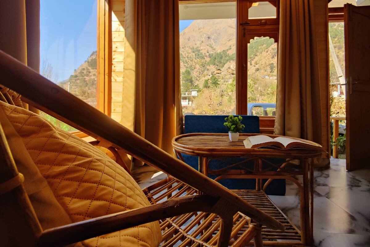A wooden-framed room with a view of mountains, a wicker chair, a small table with a plant, and an open book.