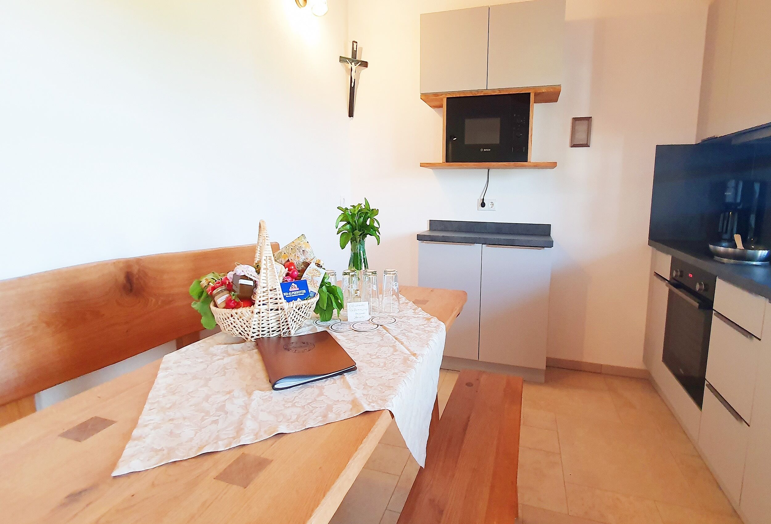 Una cocina pequeña y moderna con microondas, horno y televisor. Hay un banco de madera con una mesa que tiene un cesto de aperitivos y una planta.