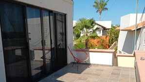 Un balcón con una silla, piso de baldosas y vistas a casas vecinas y palmeras.