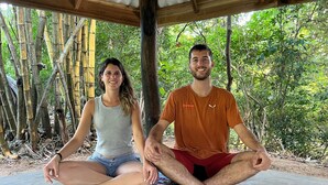 Two people sitting cross-legged on a mat under a wooden structure with bamboo and trees in the background. - River Cottage Wellawaya (Wellawaya)