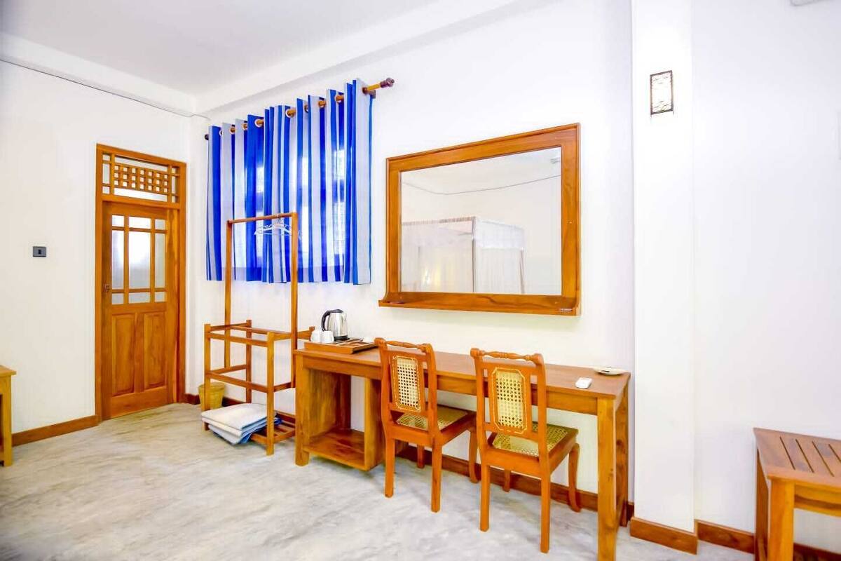 A room with a wooden desk, two chairs, a window with blue curtains, a wooden door, and a mirror on the wall.