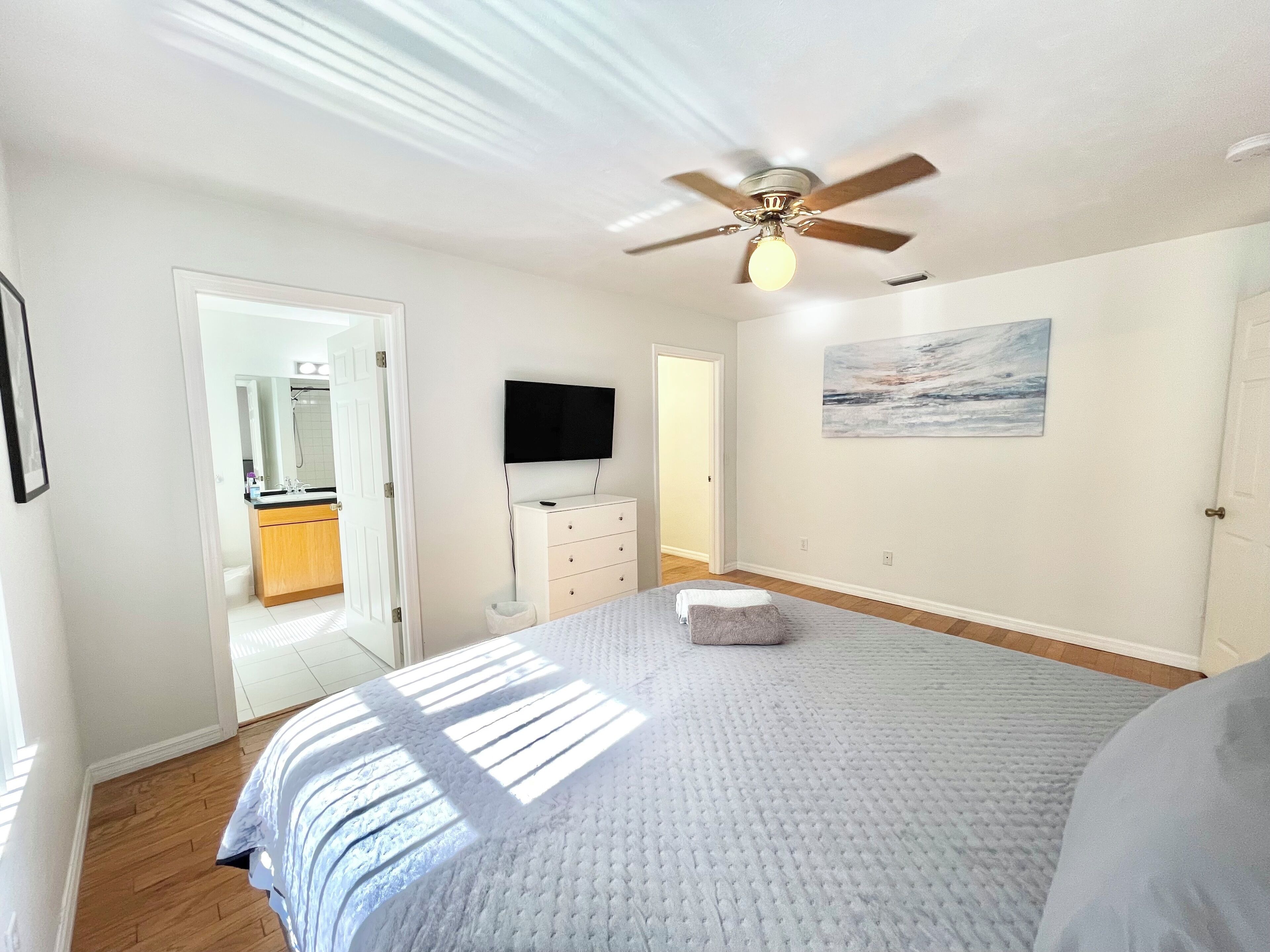 Une chambre à coucher avec un lit, un ventilateur de plafond, un téléviseur posé sur une commode et un tableau accroché au mur.