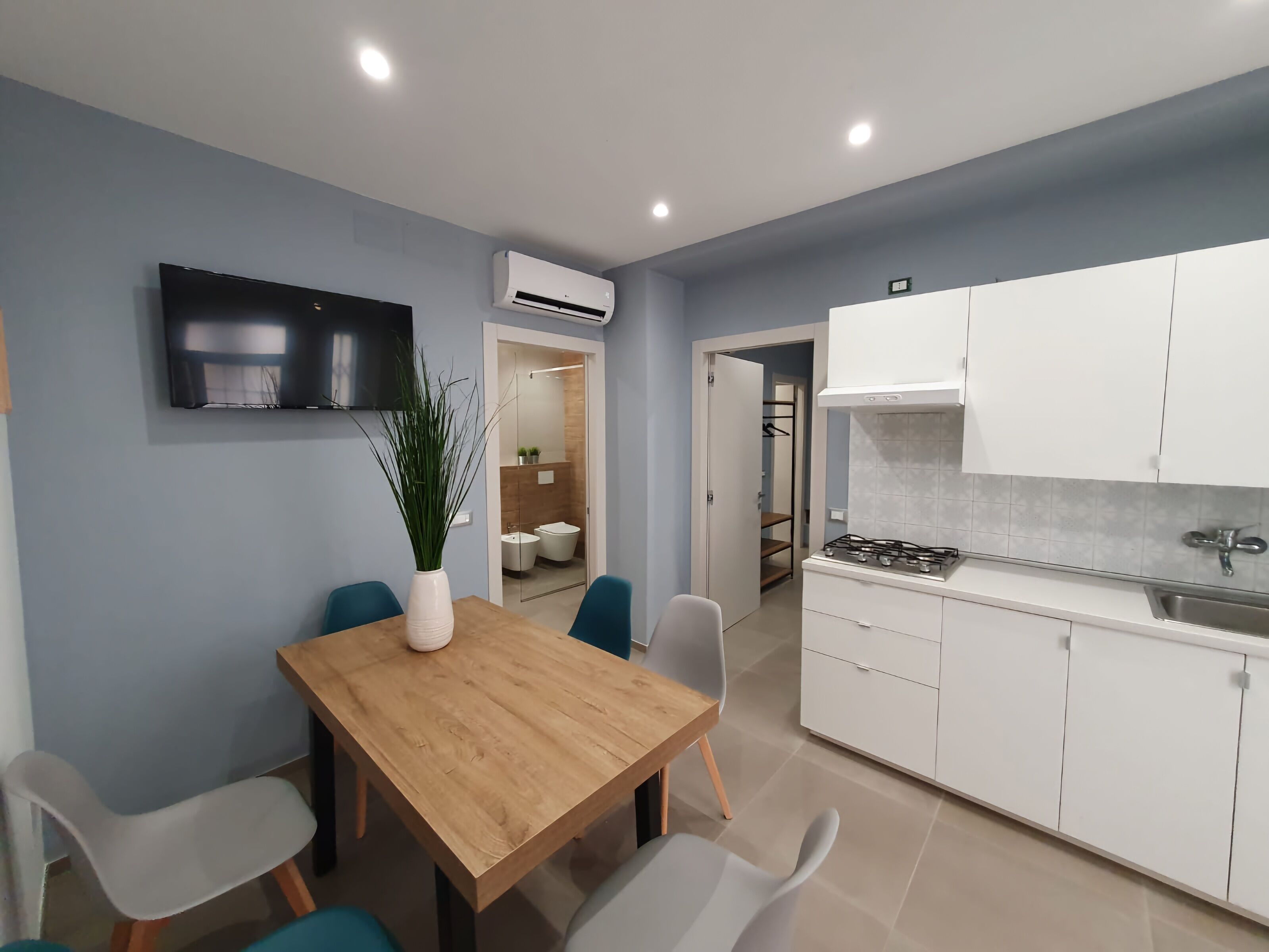 Une cuisine moderne avec des armoires blanches, une cuisinière et un évier. On y trouve également un coin repas avec une table et des chaises en bois. Un climatiseur est fixé au mur.