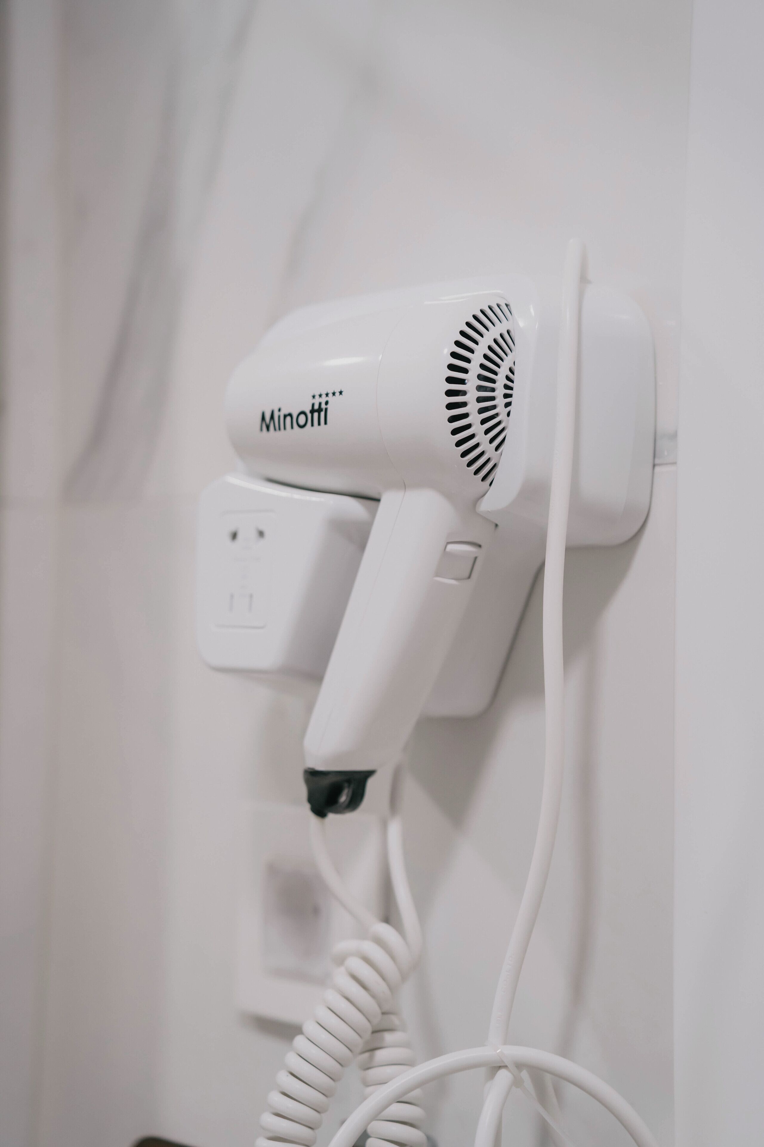 A white hair dryer mounted on a wall with a coiled cord.