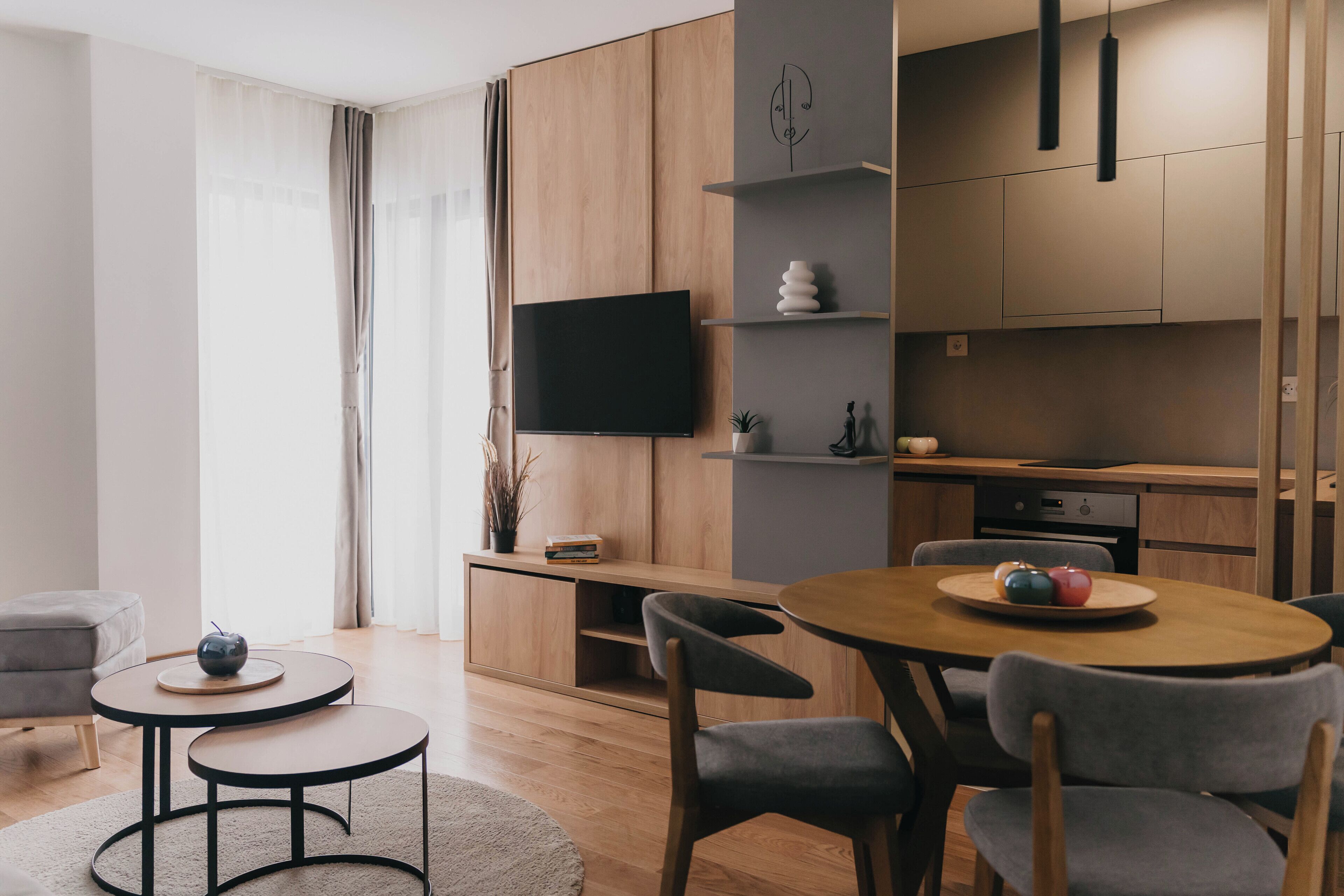 A modern living room with a wooden dining table, grey chairs, a flat-screen TV mounted on a wooden cabinet, and a small side table with a decorative bowl.