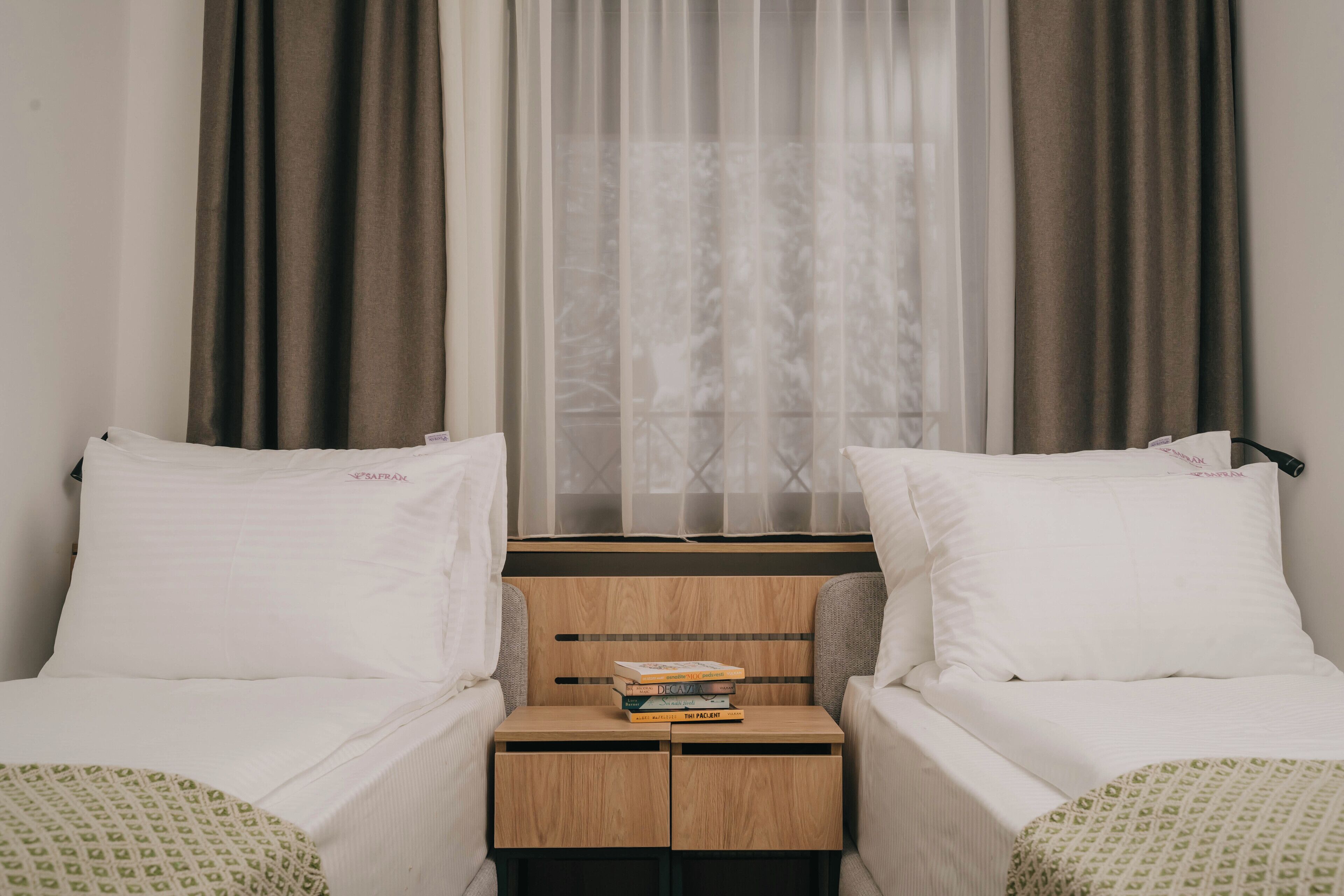 Habitación de hotel con dos camas individuales, una mesita de noche de madera con libros y cortinas traslúcidas.