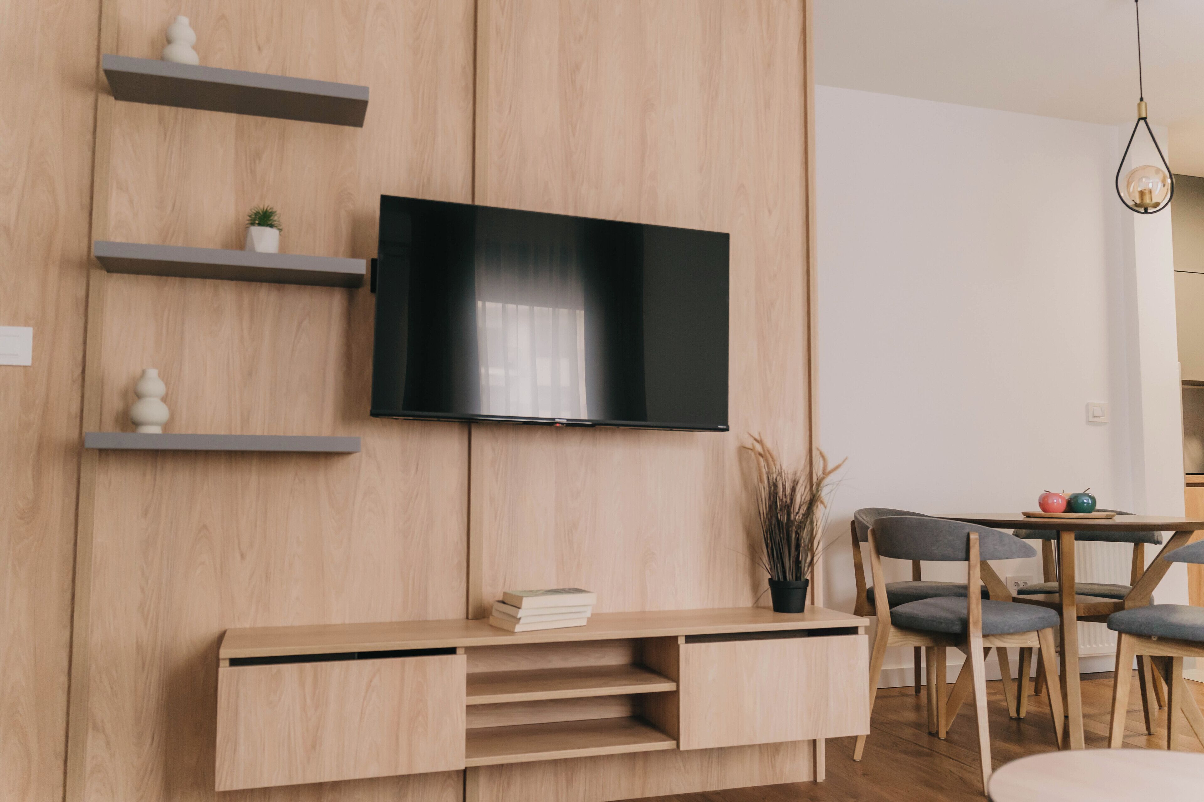 A modern living room with a flat-screen TV mounted on a wooden wall, a wooden console table with books, and a dining area with a table and chairs.
