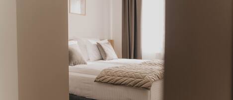Un dormitorio con cama, almohadas, una colcha y una ventana con cortinas.