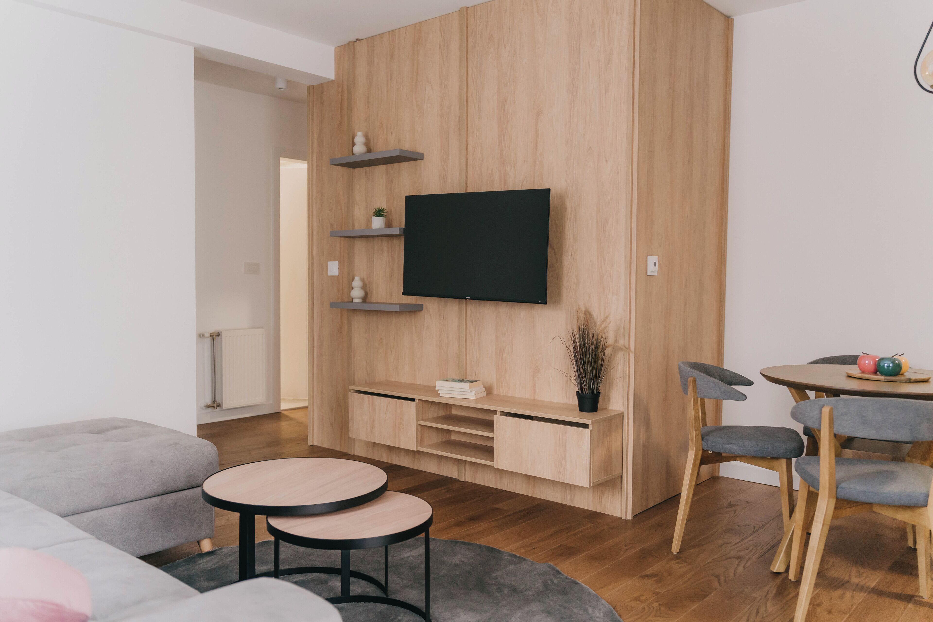 A modern living room with a flat-screen TV mounted on a wooden wall, a grey sofa, a round coffee table, and a dining area with wooden chairs and a small table.