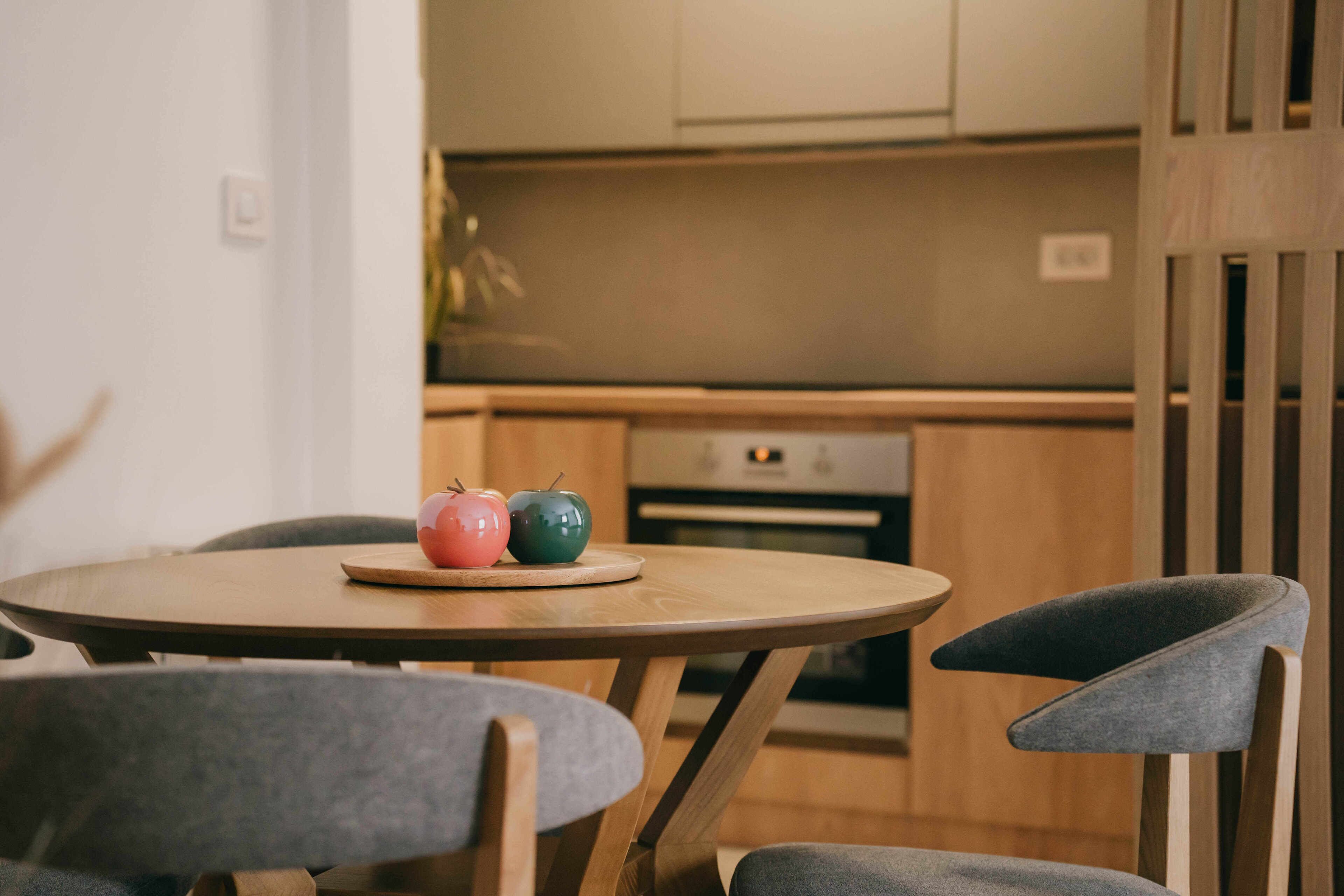 A round wooden table with two chairs, a kitchen in the background, and a cabinet above the counter.