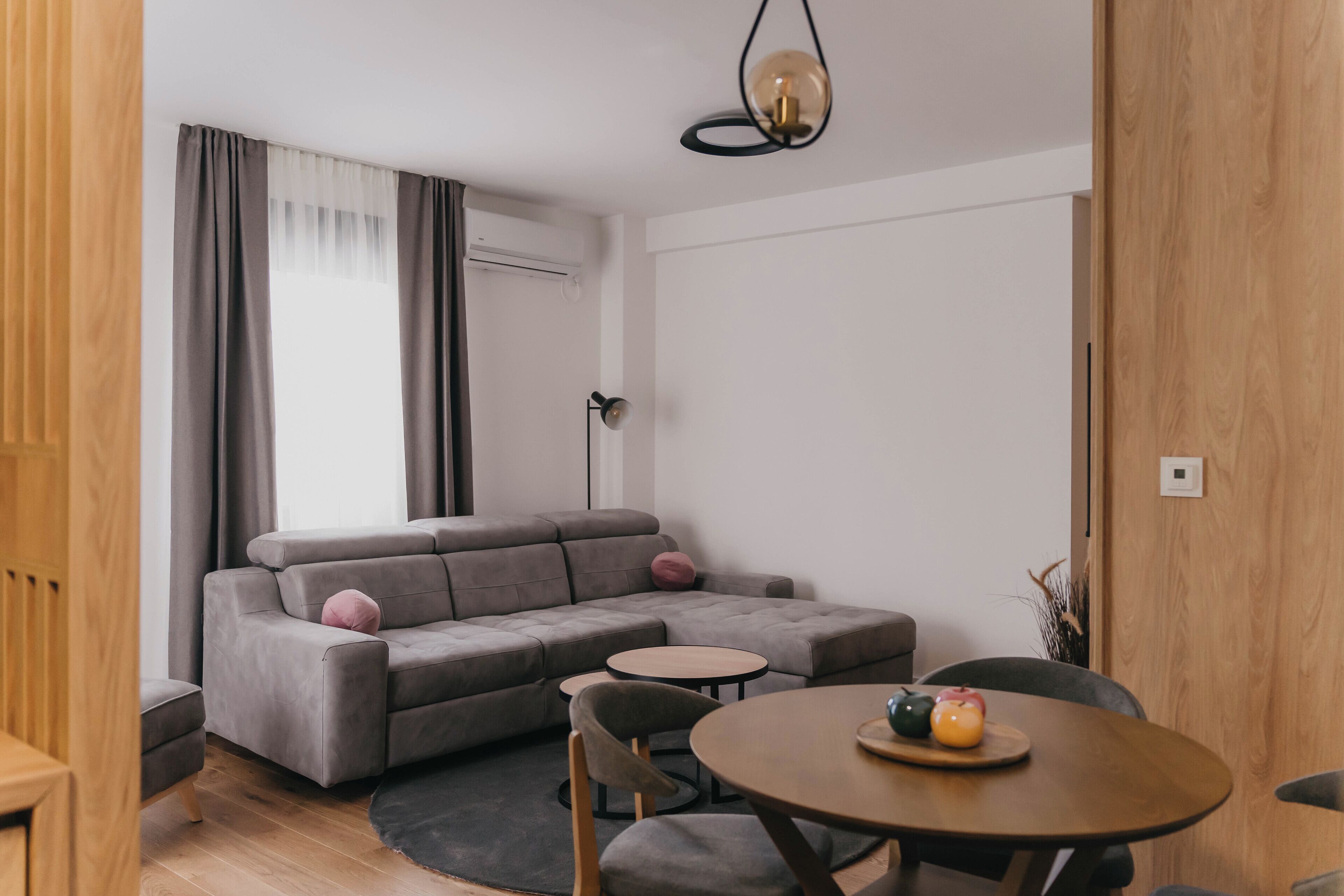 A modern living room with a grey sectional sofa, a round wooden table with a fruit bowl, and a small round table.