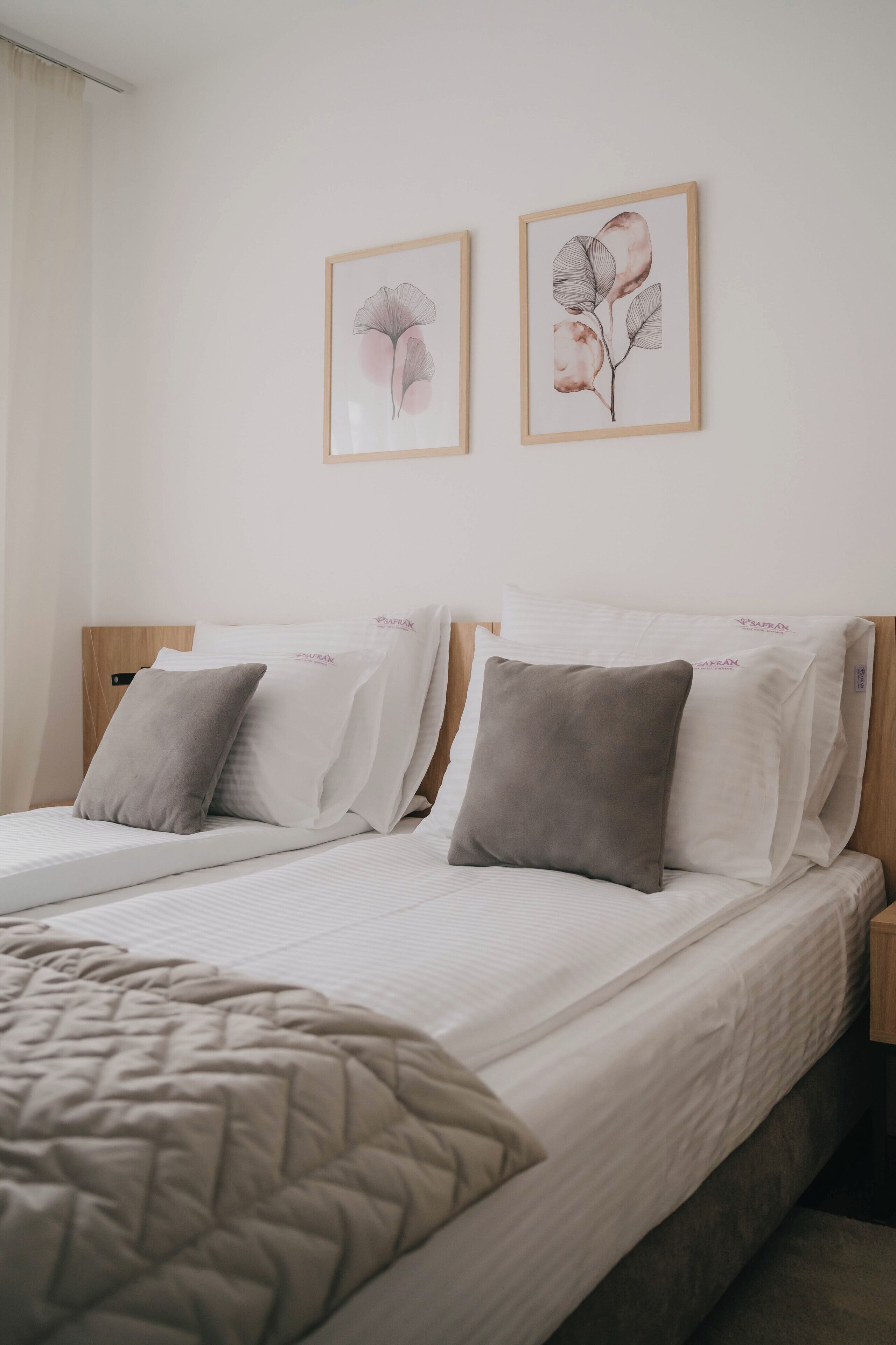 Habitación de hotel con dos camas, cada una con cabecera de madera, ropa blanca y una manta color beige. Dos láminas botánicas enmarcadas adornan la pared sobre las camas.