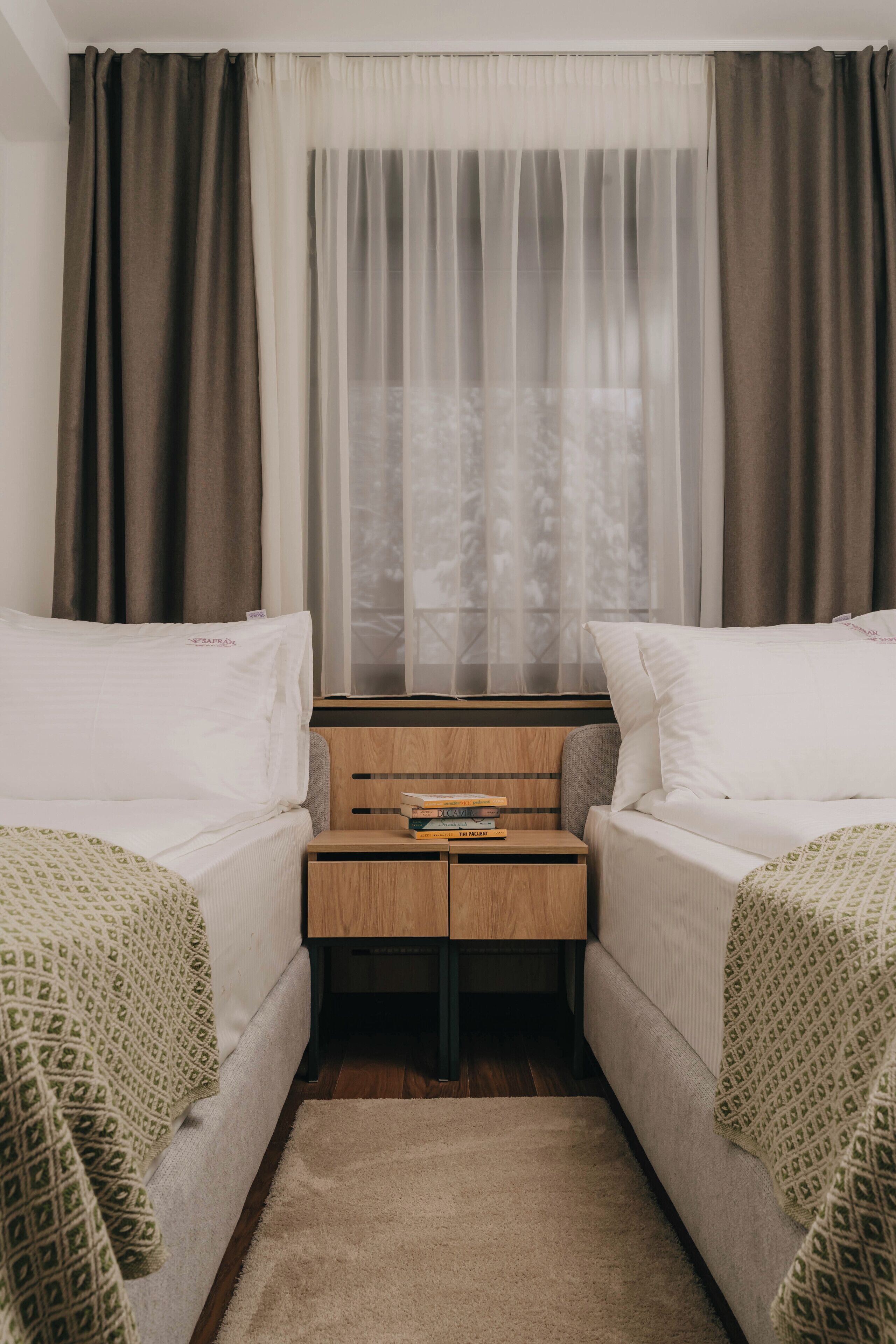 Une petite chambre à coucher, bien rangée, avec deux lits simples, une table de chevet avec des livres et une fenêtre ornée de rideaux légers.