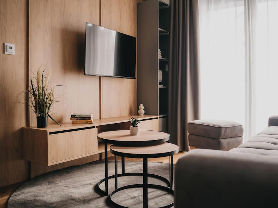 A modern, compact living space with a flat-screen TV mounted on the wall, a wooden cabinet, a small round table, a grey rug, and a grey sofa.