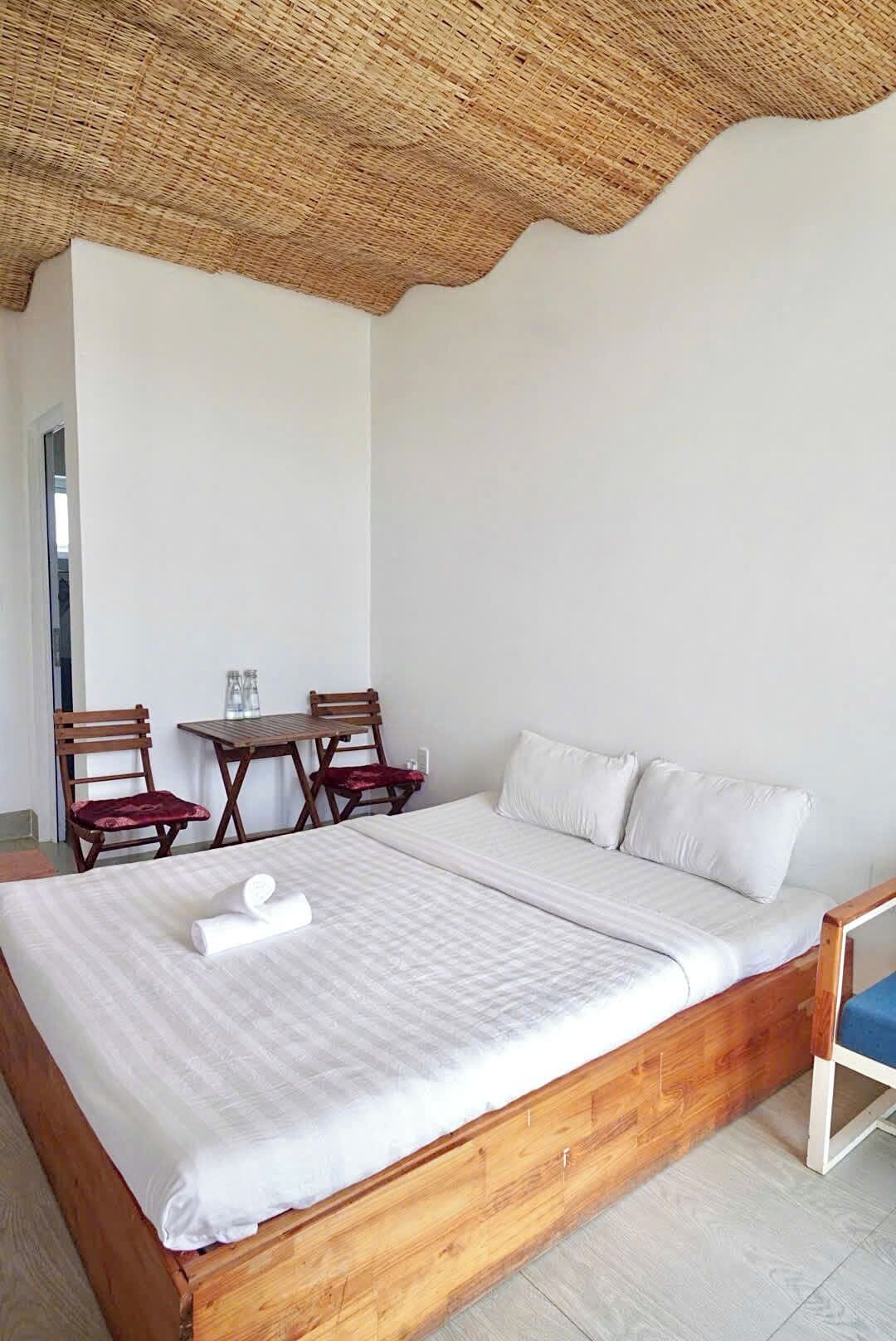 Un dormitorio con una cama de madera, ropa de cama blanca, dos sillas de madera, una mesita pequeña y un techo tejido.