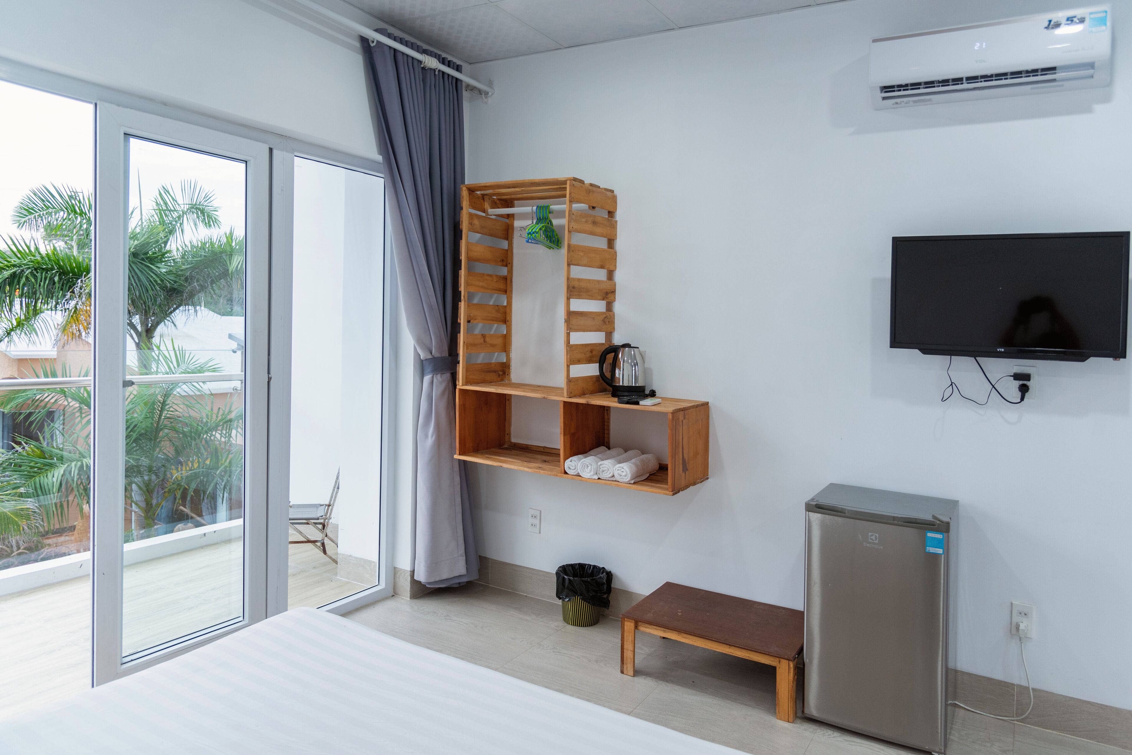 Una habitación de hotel moderna con un amplio ventanal corredero a un balcón, una estantería de madera, un televisor y un refrigerador compacto.