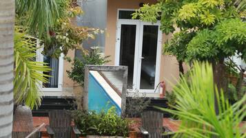 Una piscina rodeada de plantas en macetas y un edificio con fachada color crema y una ventana.