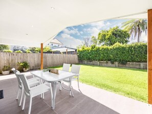 Une terrasse extérieure avec une table et des chaises, une clÎture en bois et une pelouse verdoyante.