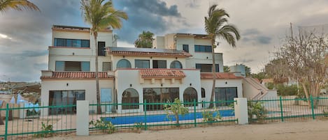 A multi-story building with a flat roof, large windows, and a blue swimming pool in the foreground.