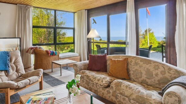 Ein gemütliches Wohnzimmer mit Sofa, Sessel und Couchtisch, großen Fenstern mit Blick auf Grünflächen und einer Flagge, sowie einer Holzbalkendecke.