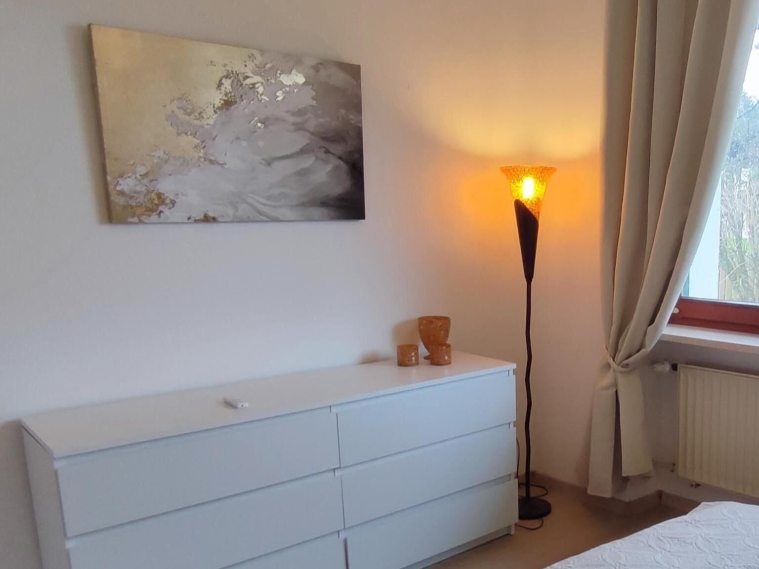 A bedroom with a white dresser, a floor lamp, and a painting on the wall.