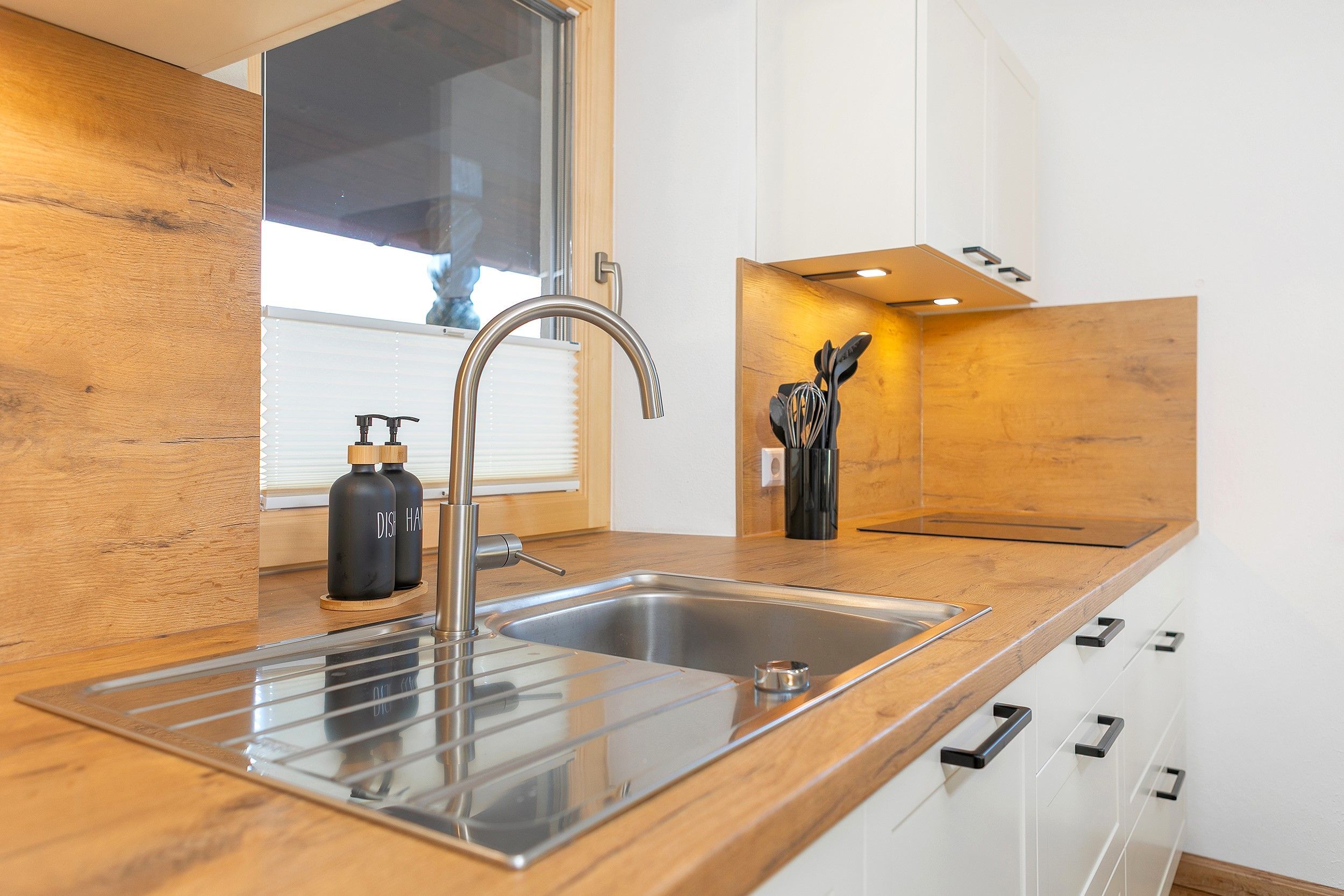 A modern kitchen with a stainless steel sink, wooden countertop, and white cabinets.