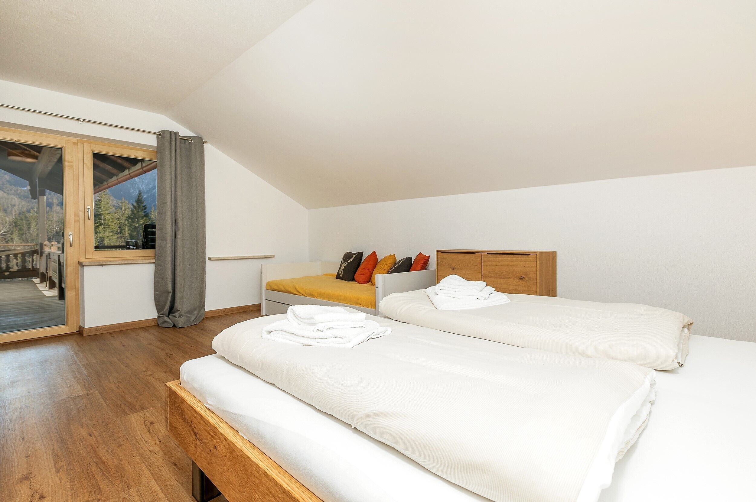 Une chambre avec deux lits, un banc en bois, une petite table et une grande fenêtre avec des rideaux.
