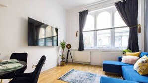 A modern living room with a blue sofa, a dining table with chairs, a mounted TV, and a large window with curtains.