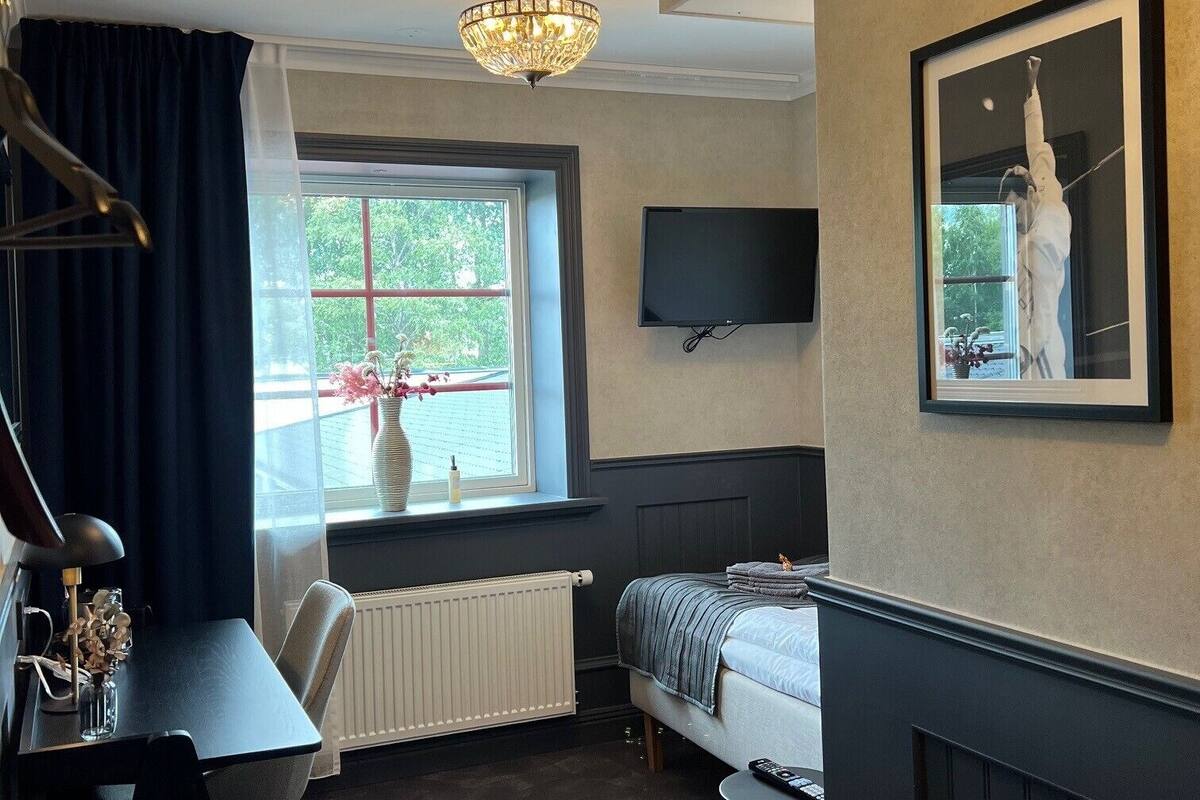 A room with a bed, a desk, a chair, a radiator, a window with curtains, a TV mounted on the wall, and a chandelier.