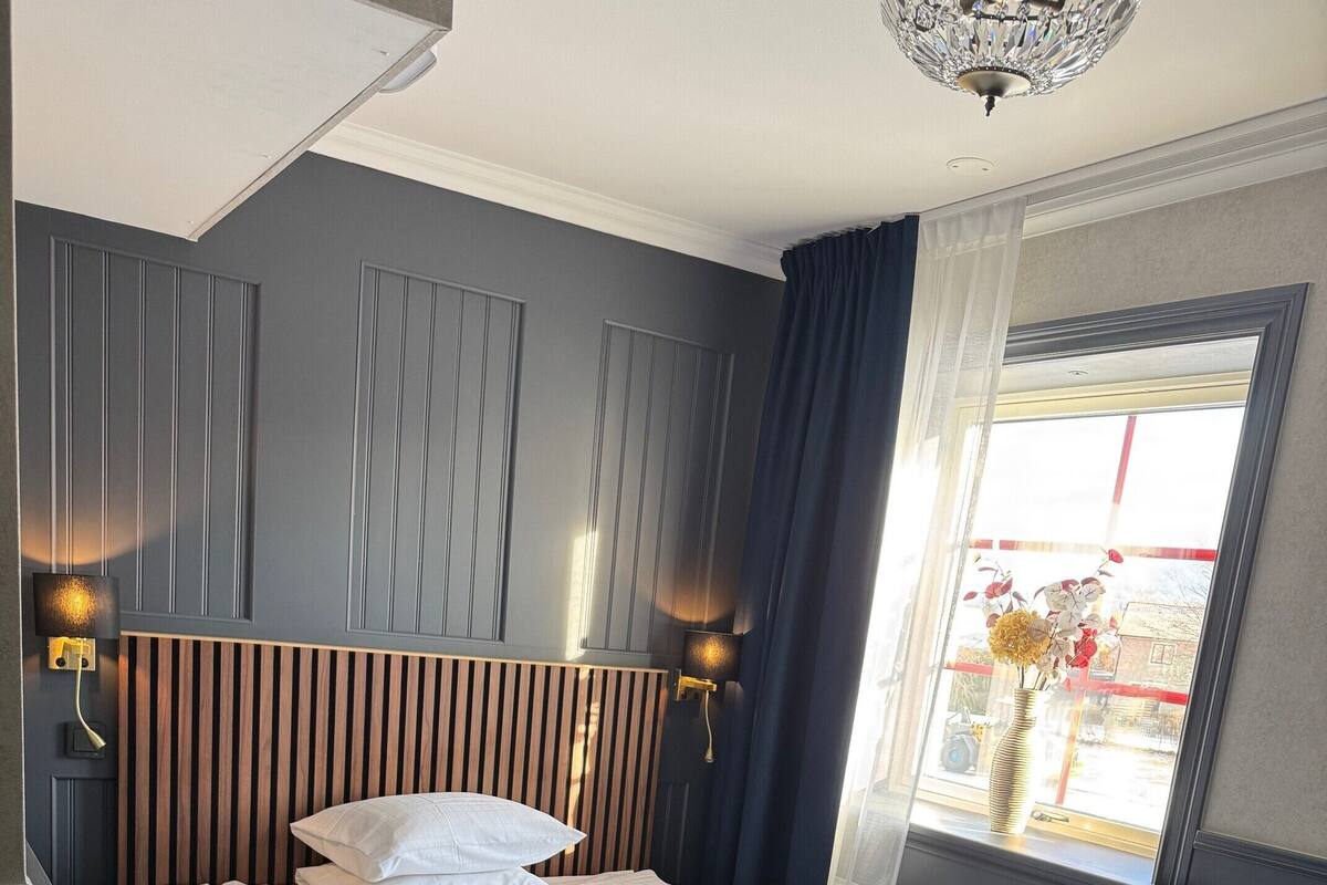 A neatly made bed with a striped blanket and pillows, a wooden headboard, two wall-mounted lamps, and a window with a view of the outside.