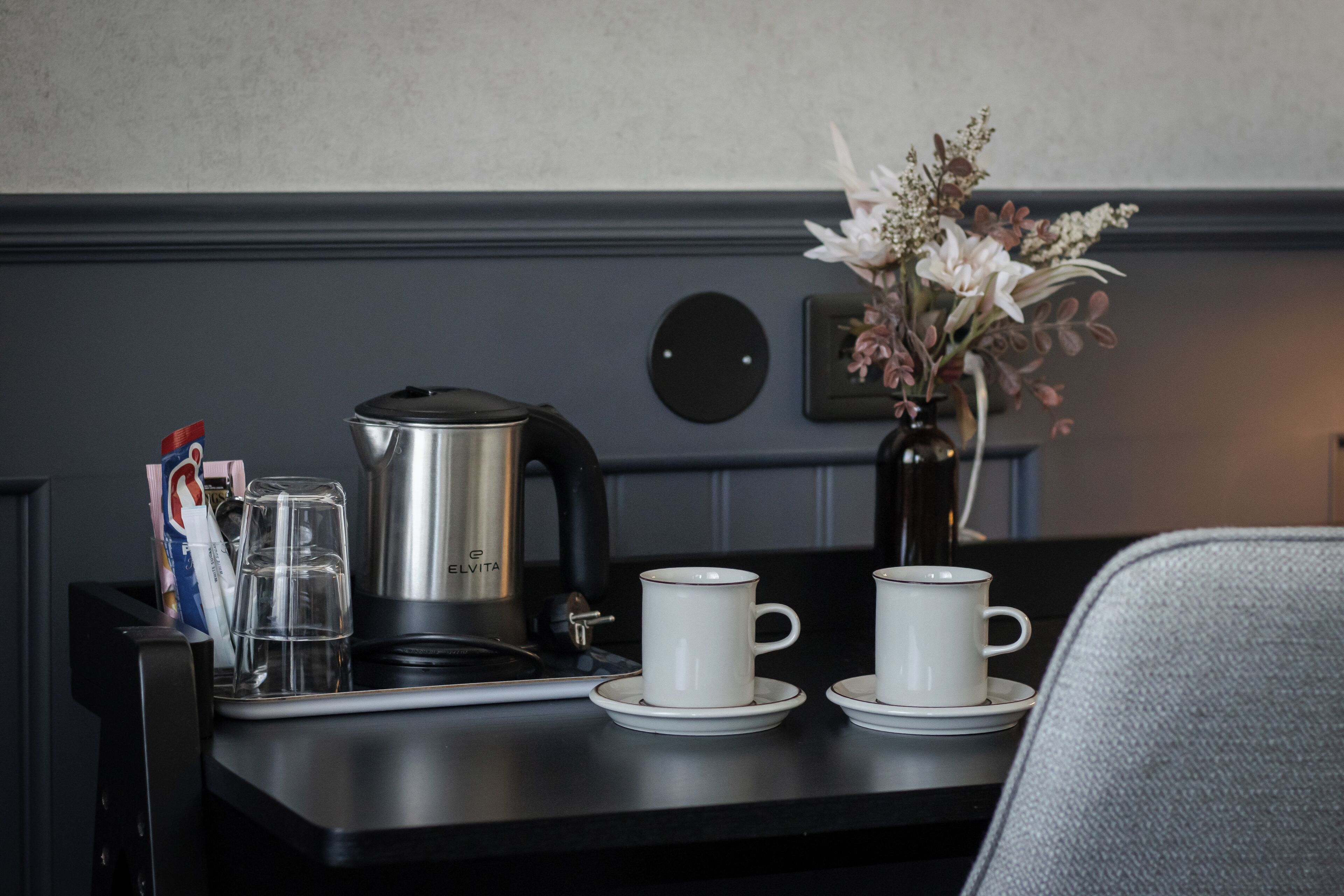 Une table mise pour le petit-déjeuner, avec une machine à café, deux tasses, un vase de fleurs et un verre d’eau.