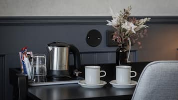 Ein Frühstücksarrangement mit Kaffeemaschine, zwei Tassen, einer Vase mit Blumen und einem Glas Wasser auf einem Tisch.