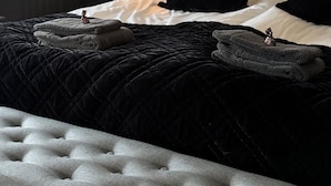 Una cama bien hecha con colcha negra, sábanas blancas y dos almohadas negras. Una lámpara de pared ilumina la habitación.