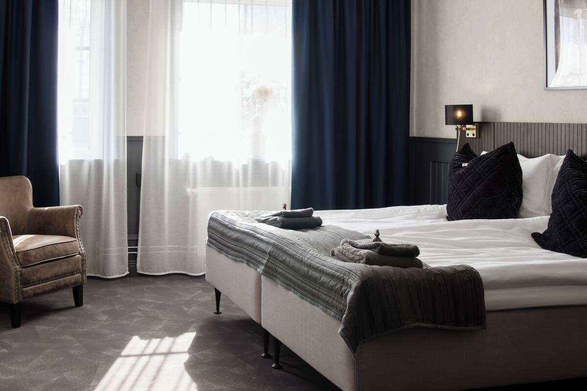 A hotel room with a bed, a chair, a bedside table with a lamp, and a window with curtains.