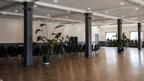Ein geräumiger Saal mit Reihen von Stühlen, großen Säulen und Kübelpflanzen.