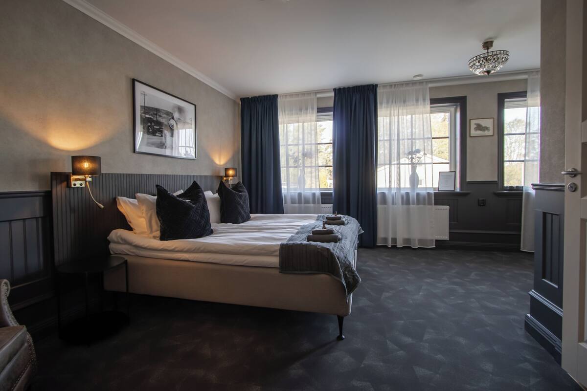 A bedroom with a large bed, two bedside tables, a chandelier, and a window with curtains.