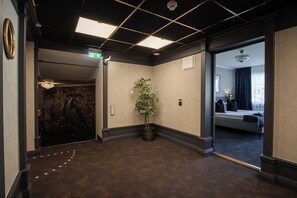 Vestíbulo de hotel con piso de madera oscura, una planta en maceta, un candelabro y una cama visible a través de una puerta abierta.