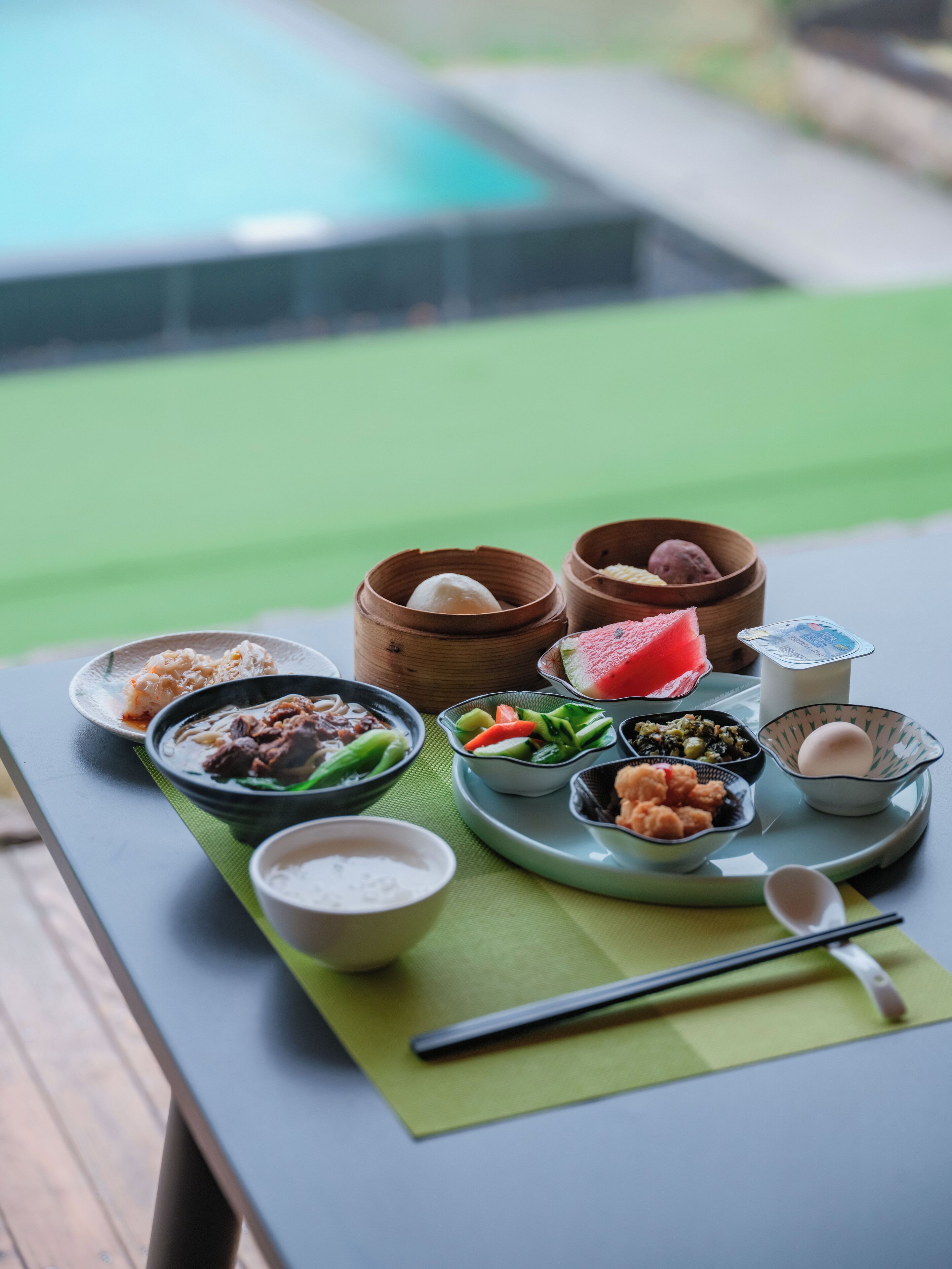 Una mesa con diversos platos, entre ellos dim sum y un tazón de sopa, ubicada al aire libre con una piscina al fondo.