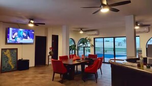 A modern dining area with a large table and red chairs, a ceiling fan, and a TV mounted on the wall.