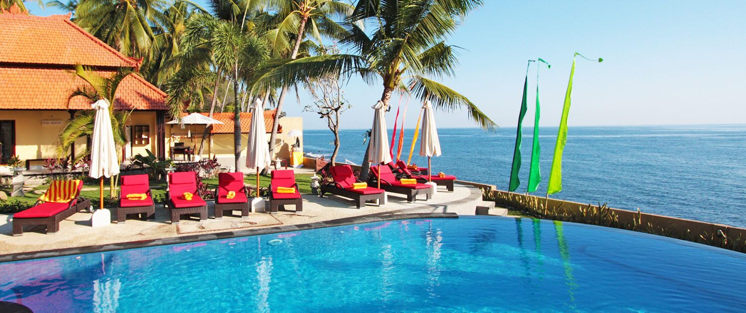A poolside area with red lounge chairs and umbrellas, a clear view of the ocean, and a building with a red-tiled roof.